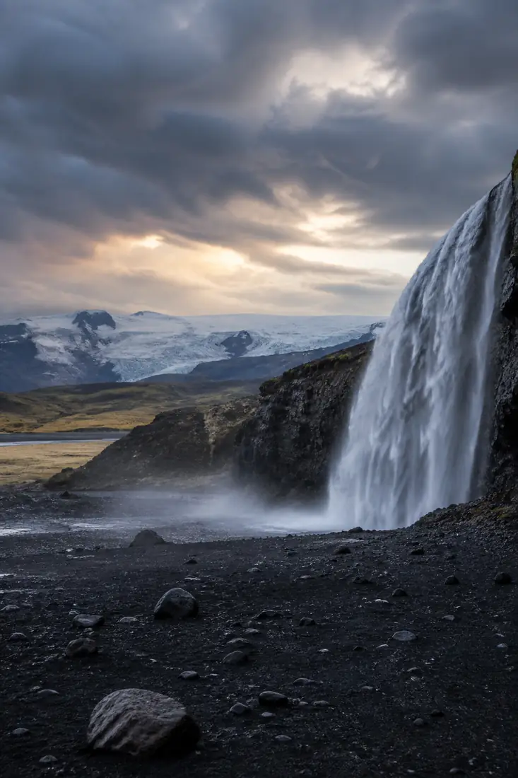 Großer Wasserfall über schwarzem Vulkansand vor einem isländischen Gletscher